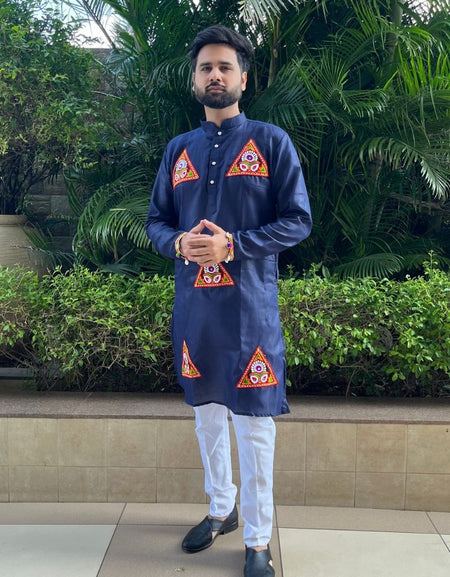 A man wearing a navy blue kurta with red and gold geometric patterns on the chest and lower hem, paired with white trousers and black shoes, standing outdoors in front of green plants.