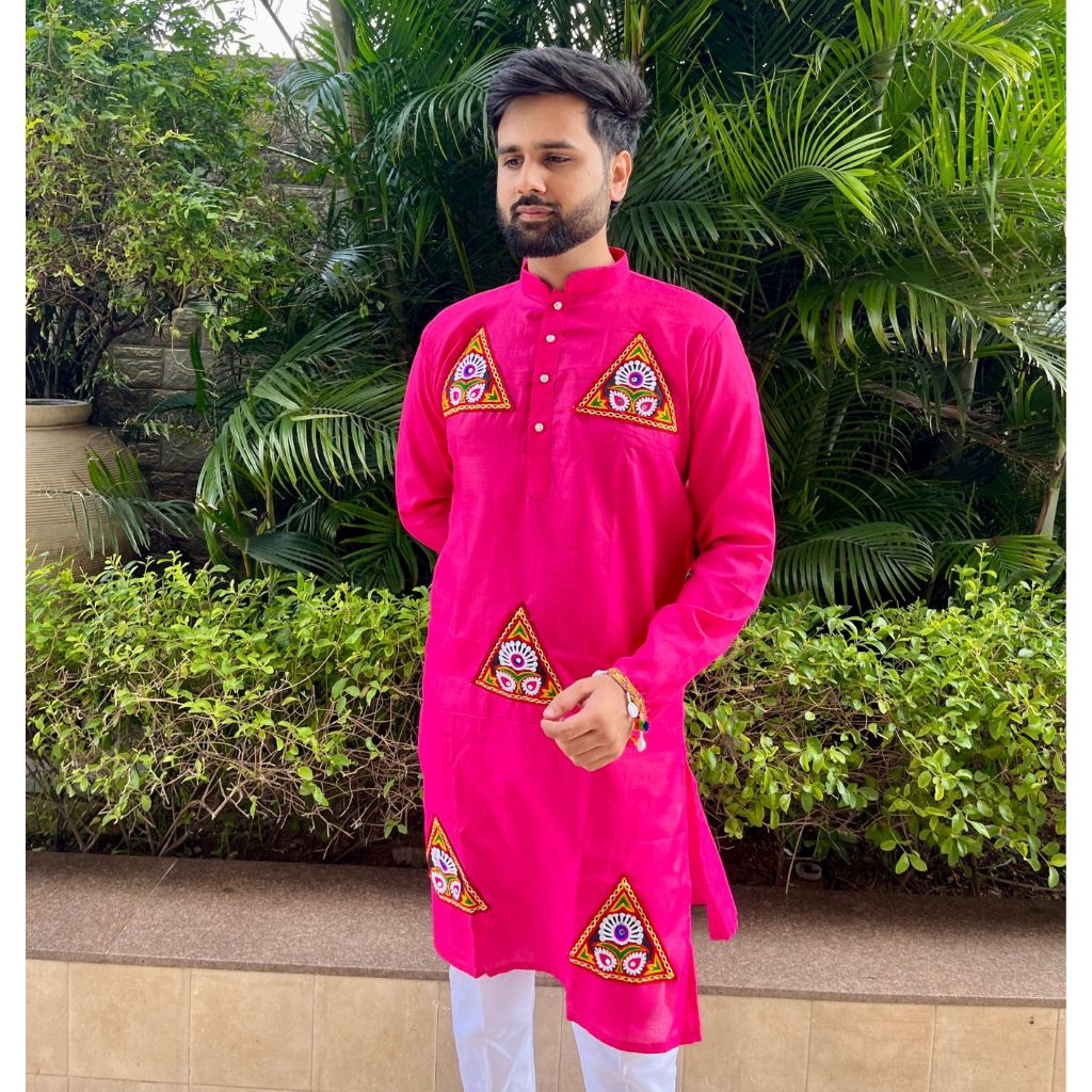 A man wearing a bright pink kurta with embroidered geometric patterns on the chest and lower hem, paired with white trousers, standing outdoors in front of green tropical foliage.
