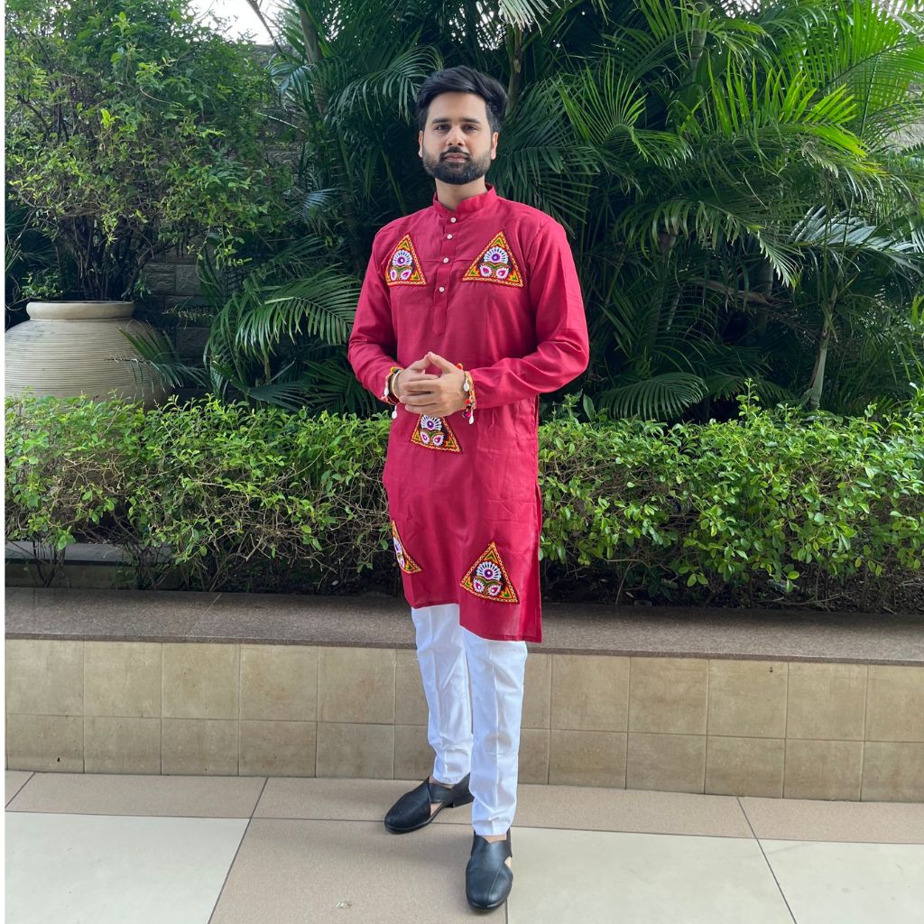 A man wearing a bright red kurta with yellow and white geometric embroidery on the chest and hem, paired with white trousers and black shoes, standing outdoors in front of green plants.