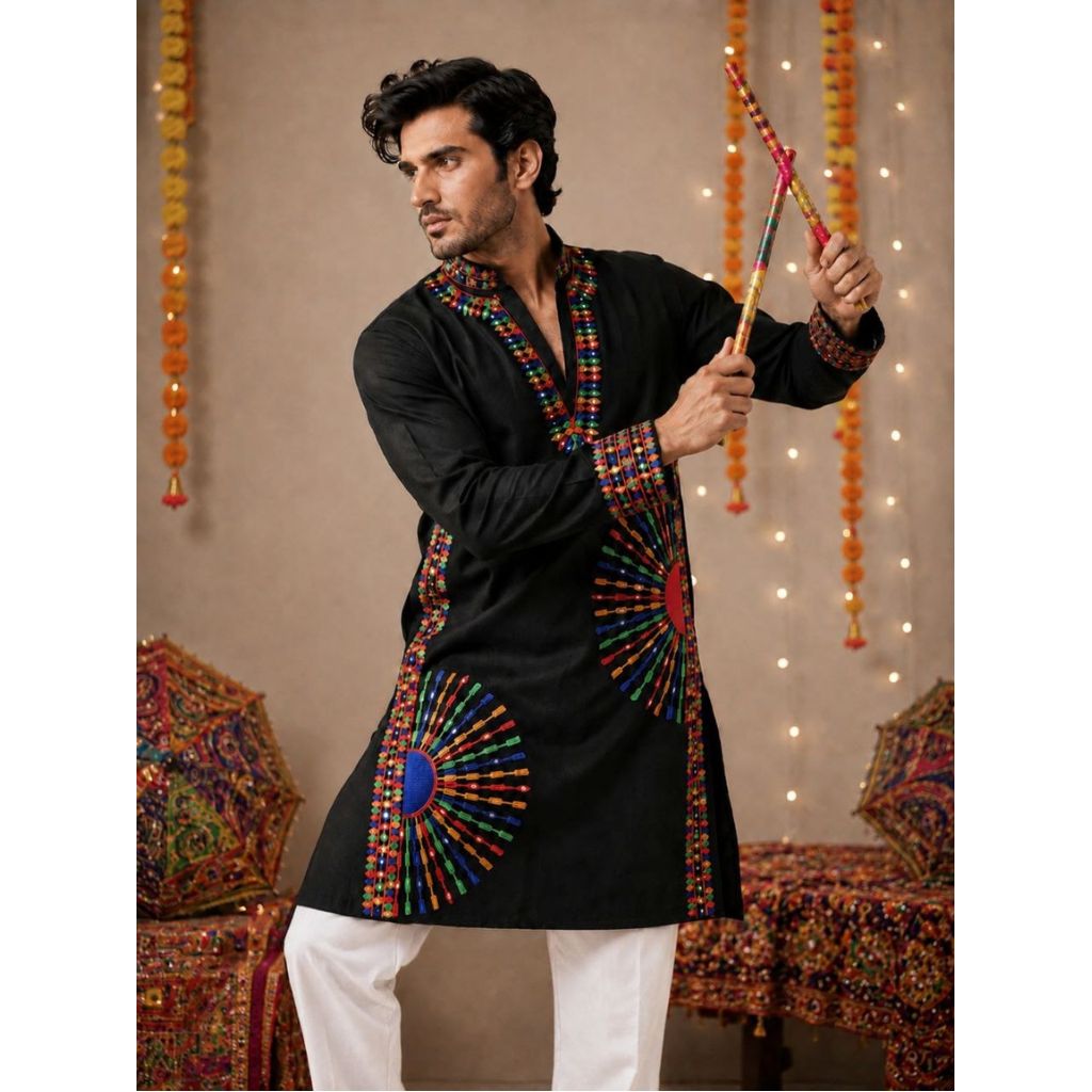 A man wearing a black kurta with colorful embroidered patterns and white pants, holding a decorative staff in a festive indoor setting with hanging marigold garlands and string lights.