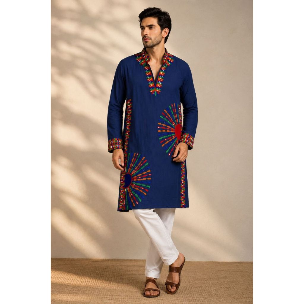 A man modeling a navy blue kurta with colorful embroidered patterns on the neckline, cuffs, and hem, paired with white trousers and brown sandals, set against a beige wall with soft shadow patterns.