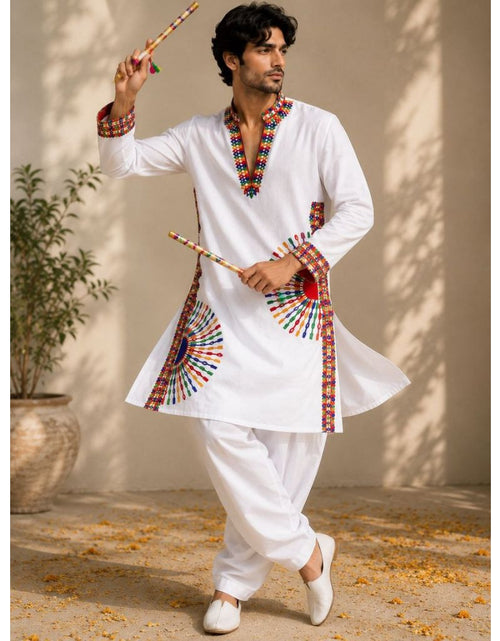 Load image into Gallery viewer, A man modeling a white traditional Indian kurta and pant set with colorful embroidered details, holding wooden sticks against a warm beige wall with leaf shadows.

