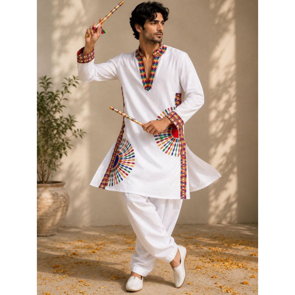 A man modeling a white traditional Indian kurta and pant set with colorful embroidered details, holding wooden sticks against a warm beige wall with leaf shadows.