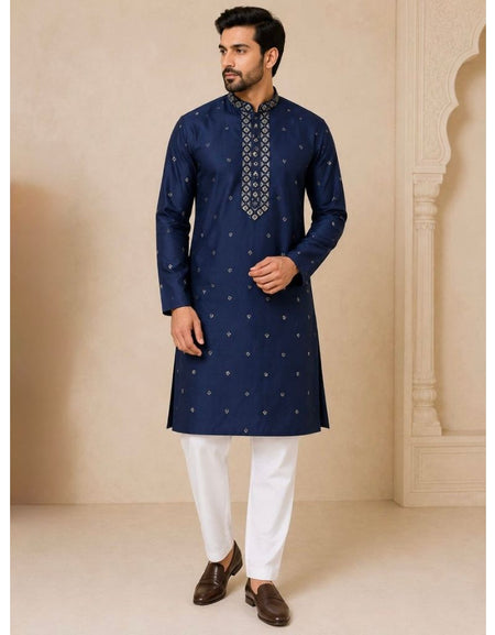 A man modeling a navy blue kurta with small gold motifs and a patterned mandarin collar, paired with white trousers and brown shoes, set against a beige architectural background.