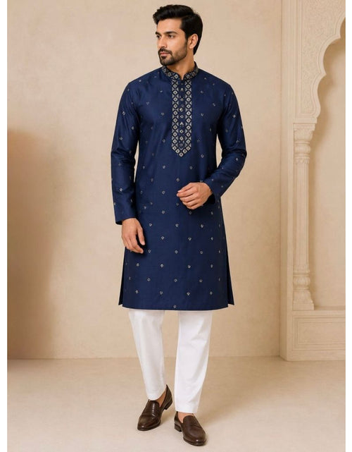 Load image into Gallery viewer, A man modeling a navy blue kurta with small gold motifs and a patterned mandarin collar, paired with white trousers and brown shoes, set against a beige architectural background.
