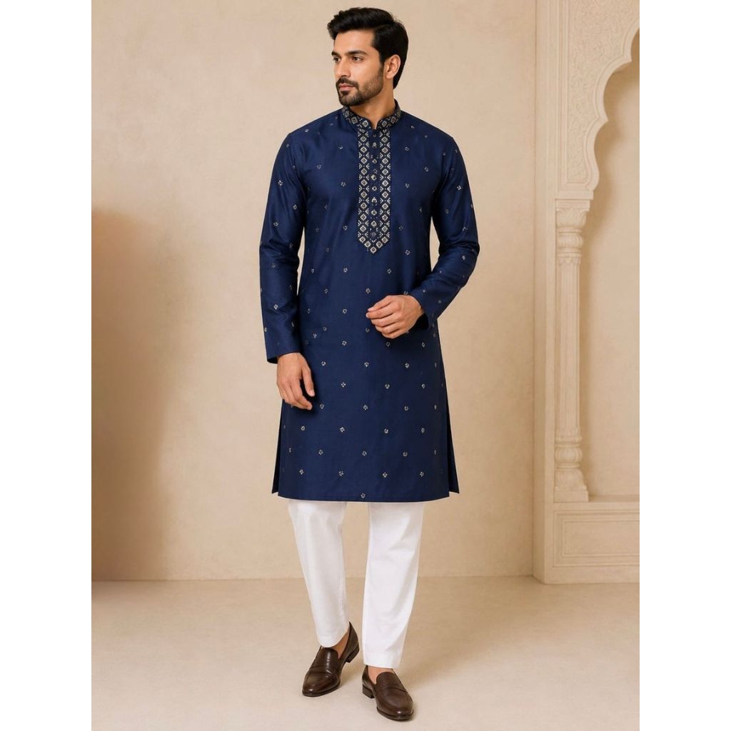 A man modeling a navy blue kurta with small gold motifs and a patterned mandarin collar, paired with white trousers and brown shoes, set against a beige architectural background.