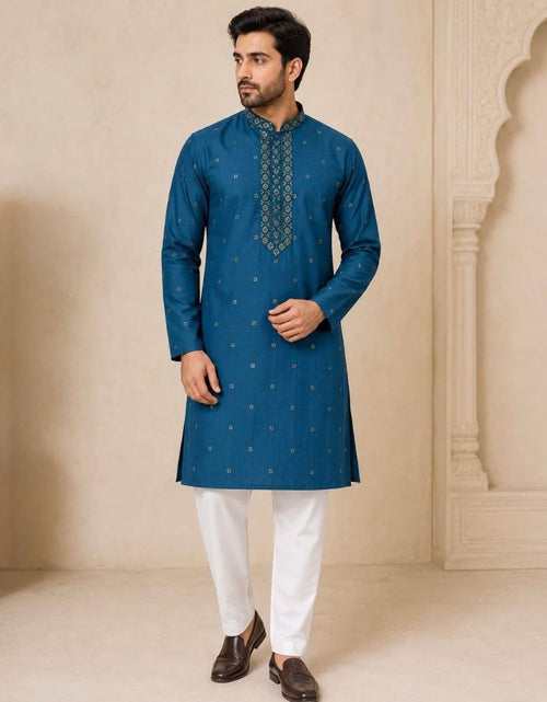 Load image into Gallery viewer, A man modeling a teal blue kurta with small gold motifs and a decorative embroidered yoke, paired with white trousers and brown shoes, standing in a beige architectural setting.
