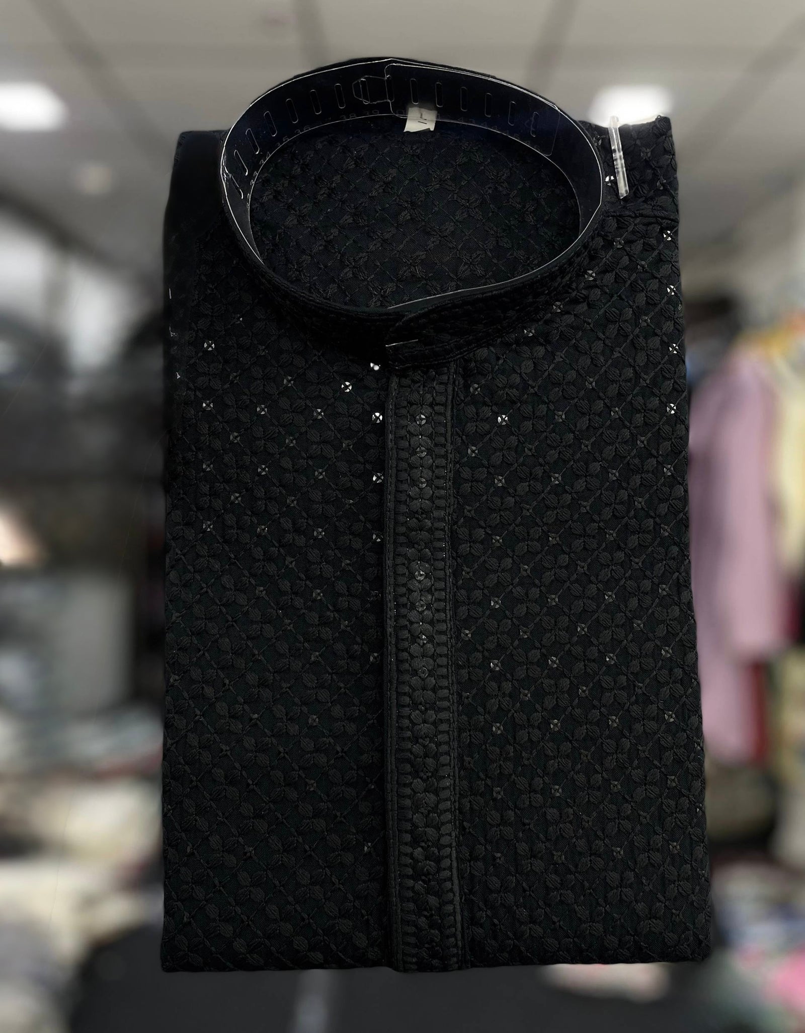 A folded black textured kurta with a stand collar and subtle silver embellishments, displayed against a blurred indoor background.