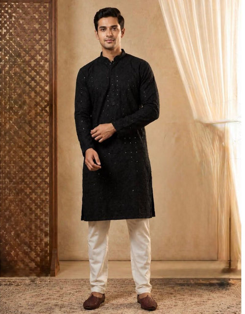 Load image into Gallery viewer, A man modeling a black long-sleeve kurta with subtle silver embroidery, paired with off-white trousers and brown loafers, standing in a warm-toned studio setting with a textured screen and draped curtains.
