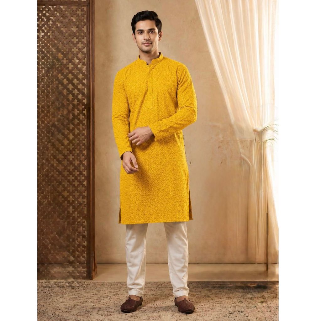 A man modeling a bright yellow long-sleeve kurta with white trousers and brown shoes, standing in a warm-toned indoor setting with patterned screens and draped curtains.