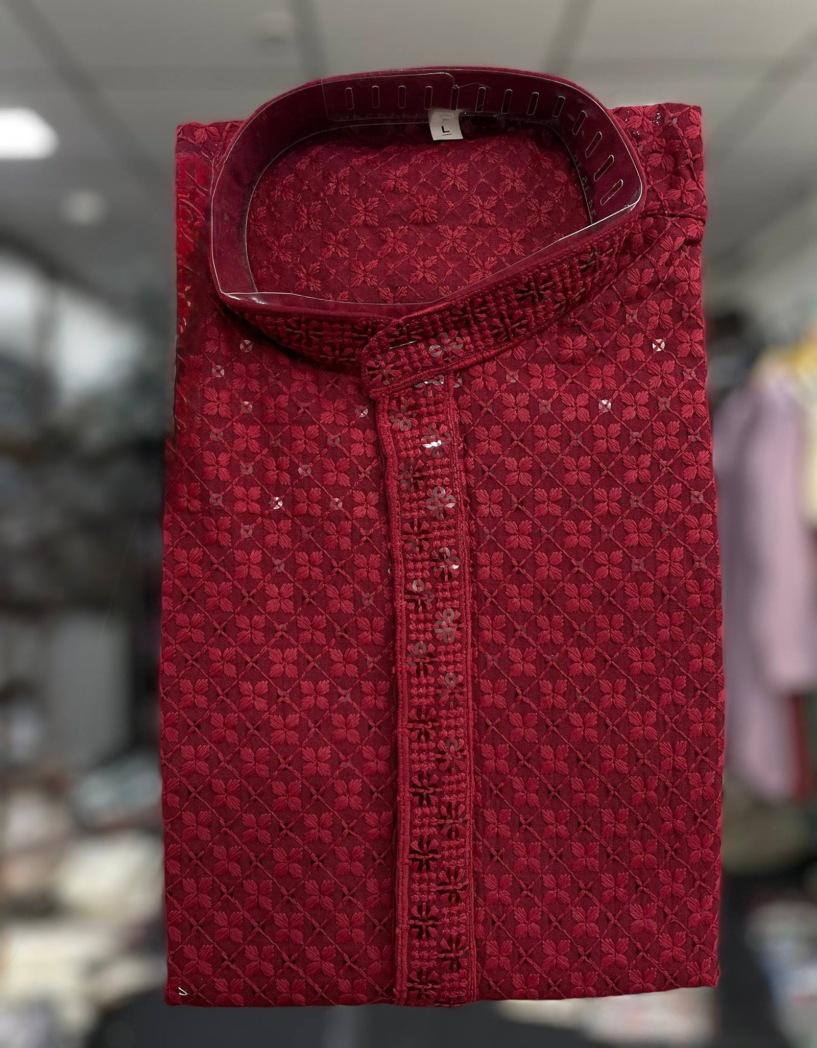 A folded maroon red kurta with a textured geometric pattern and sequined button placket, displayed against a blurred background.