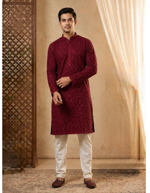 Load image into Gallery viewer, A man modeling a deep maroon long-sleeve kurta with a subtle floral pattern, paired with off-white trousers and brown shoes, standing in a warm, decorated indoor setting.
