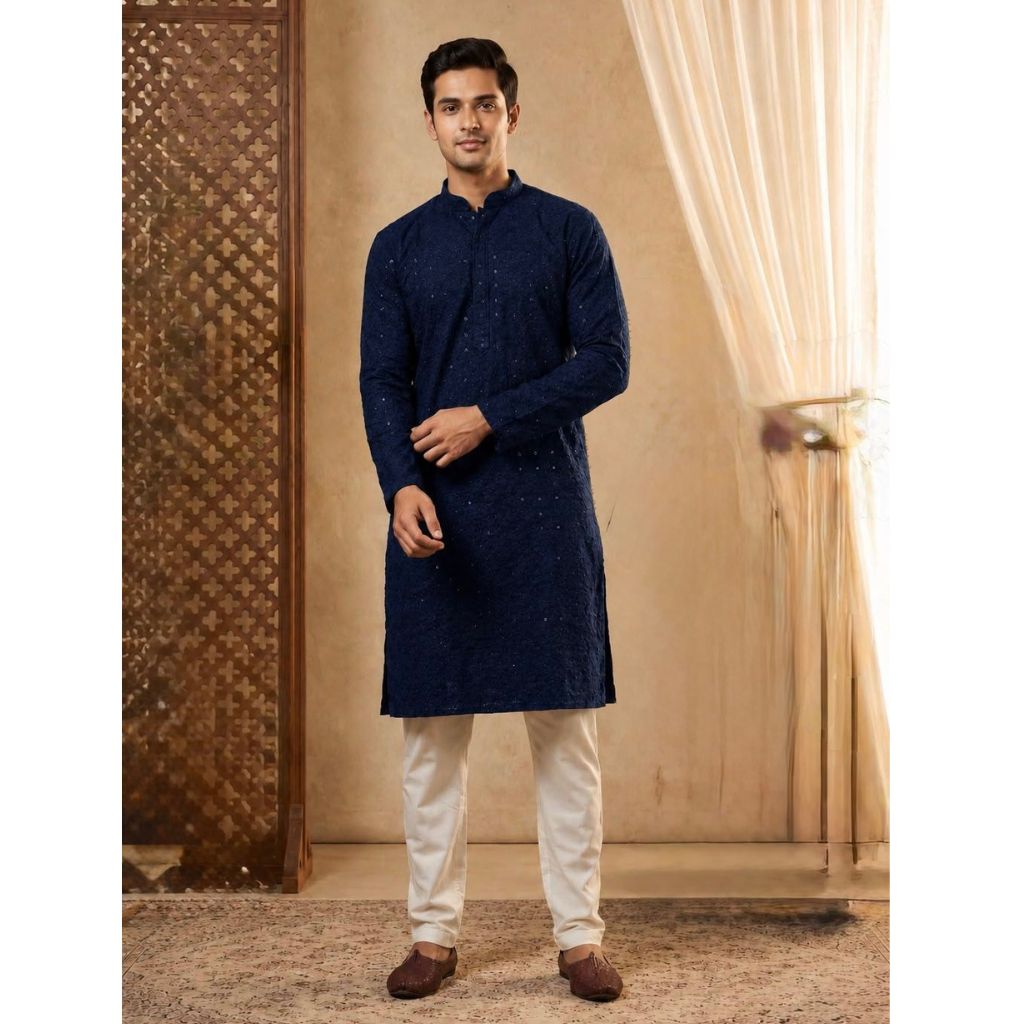 A man modeling a dark navy blue kurta with subtle embroidered patterns, paired with off-white trousers and brown shoes, standing in a warm-toned indoor setting with curtains.