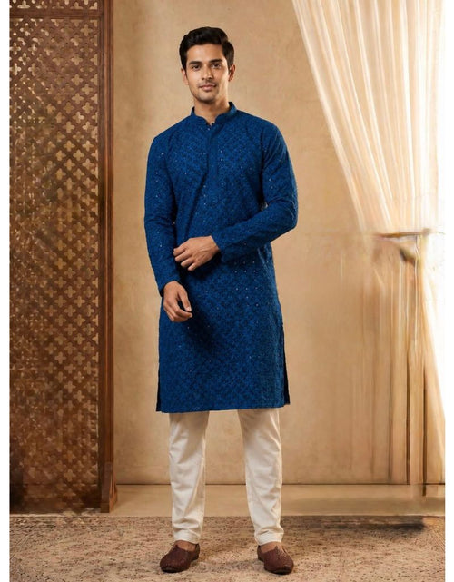 Load image into Gallery viewer, A man modeling a royal blue patterned kurta with cream trousers and brown shoes, standing in a warm-toned indoor setting with a patterned screen and sheer curtains.
