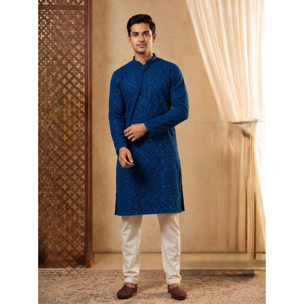 A man modeling a royal blue patterned kurta with cream trousers and brown shoes, standing in a warm-toned indoor setting with a patterned screen and sheer curtains.