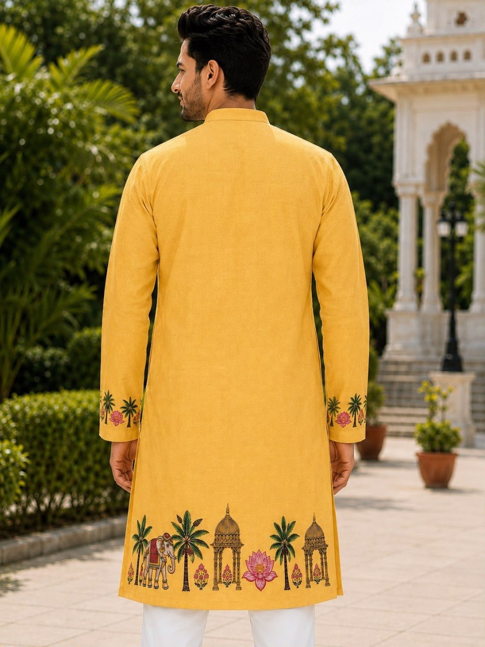 A man viewed from behind wearing a bright yellow kurta with colorful embroidered motifs of palm trees, domes, and lotus flowers near the hem and cuffs, set against a garden background with a white pavilion.