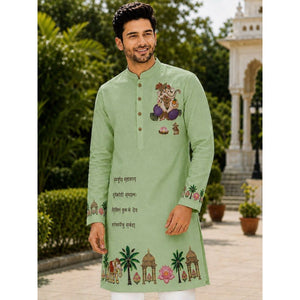 A man wearing a light green long-sleeve kurta with colorful traditional Indian illustrations and text on the chest and cuffs, standing outdoors in front of a garden and white architectural structure.