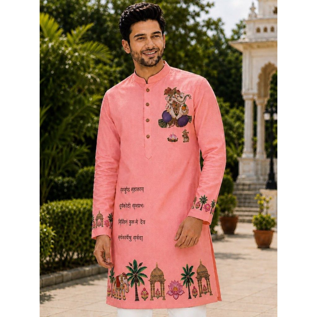 A man wearing a pink Indian kurta with traditional embroidery and text, standing outdoors in front of a garden and architectural background.