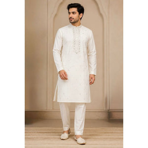 A man wearing a traditional white Indian kurta and matching trousers, standing in front of a beige arched wall background.