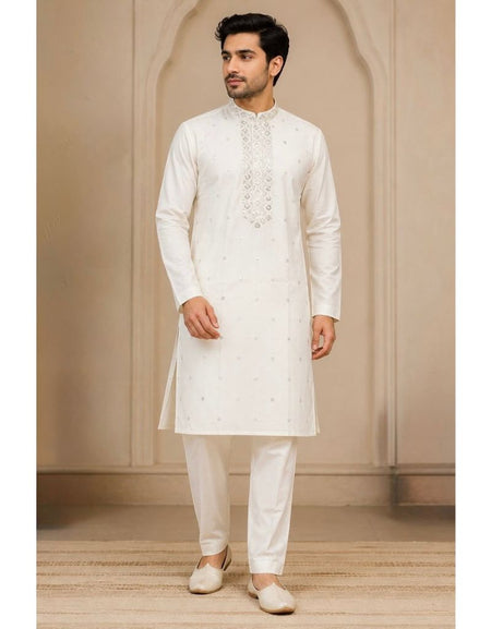 A man wearing a traditional white Indian kurta and matching trousers, standing in front of a beige arched wall background.