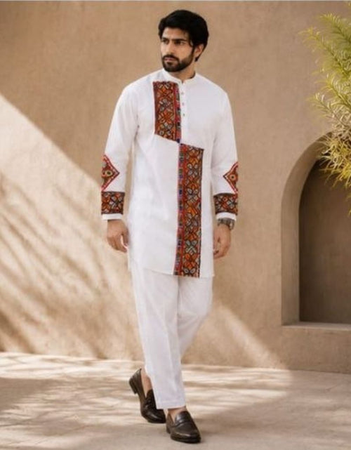 Load image into Gallery viewer, A man wearing a white traditional kurta set with red and multicolor geometric embroidery on the chest, sleeves, and front pocket, paired with white trousers and dark brown loafers, standing in front of a beige stucco wall with shadows.
