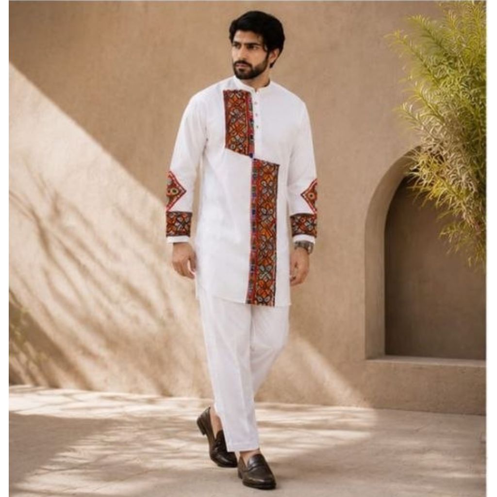 A man wearing a white traditional kurta set with red and multicolor geometric embroidery on the chest, sleeves, and front pocket, paired with white trousers and dark brown loafers, standing in front of a beige stucco wall with shadows.