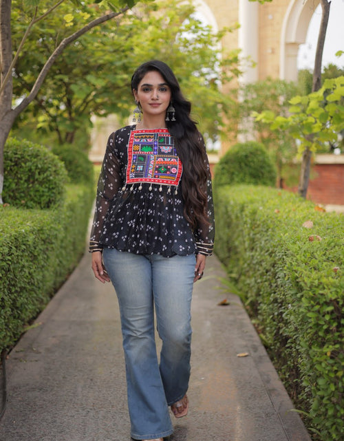 Load image into Gallery viewer, A woman walking on a paved path outdoors, wearing a black long-sleeve blouse with a colorful embroidered chest panel and light blue jeans.
