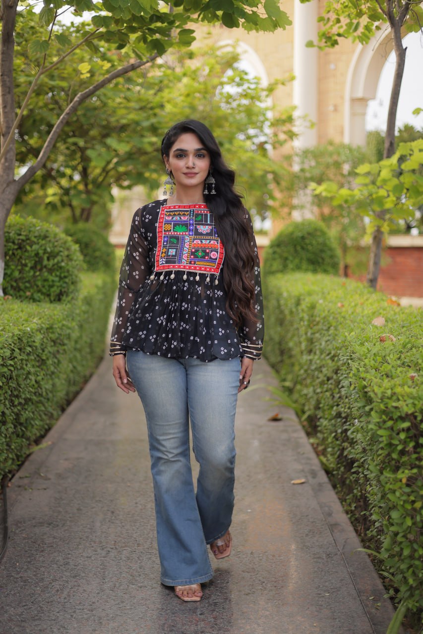A woman walking on a paved path outdoors, wearing a black long-sleeve blouse with a colorful embroidered chest panel and light blue jeans.