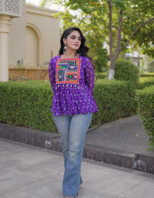 Load image into Gallery viewer, A woman wearing a purple long-sleeve embroidered top with blue jeans, standing outdoors on a paved walkway with hedges and a building in the background.
