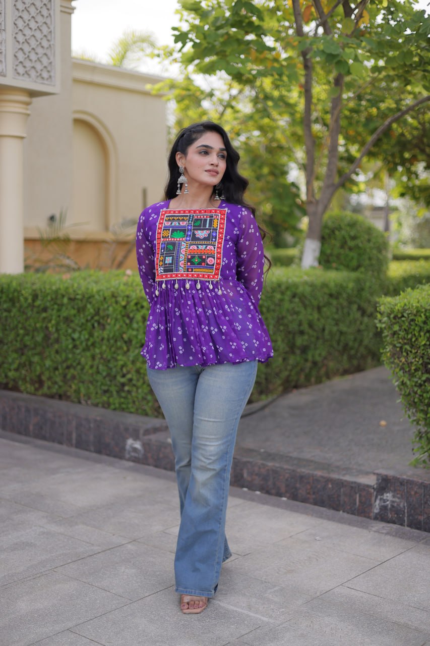 A woman wearing a purple long-sleeve embroidered top with blue jeans, standing outdoors on a paved walkway with hedges and a building in the background.