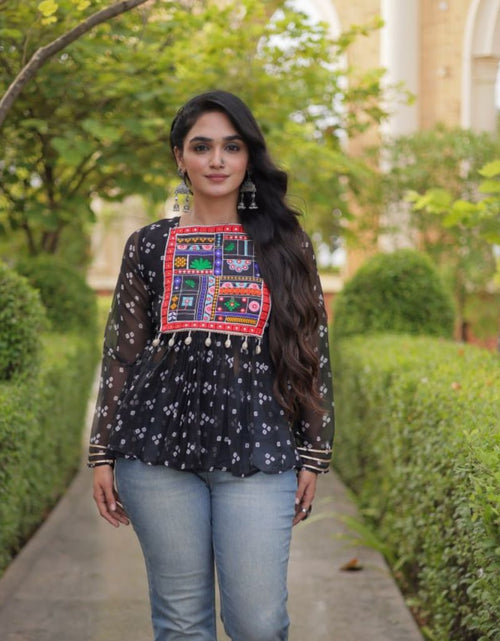 Load image into Gallery viewer, A woman stands on a garden path wearing a black long-sleeve blouse with white floral embroidery and a colorful embroidered chest panel, paired with light blue jeans.
