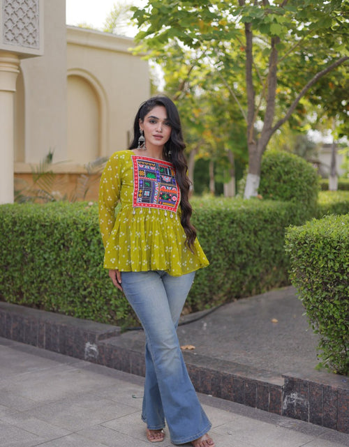 Load image into Gallery viewer, A woman stands outdoors on a paved walkway wearing a bright yellow-green embroidered long-sleeve top with blue flared jeans. The background features trimmed green hedges, trees, and a beige building.
