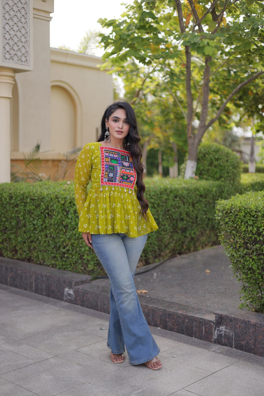 A woman stands outdoors on a paved walkway wearing a bright yellow-green embroidered long-sleeve top with blue flared jeans. The background features trimmed green hedges, trees, and a beige building.
