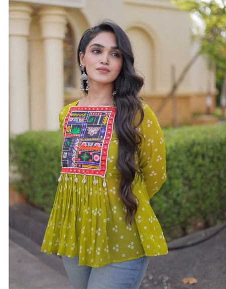 A woman wearing a yellow-green ethnic kurta with a colorful embroidered patch on the chest, paired with light blue jeans, standing outdoors in front of a beige building with columns.
