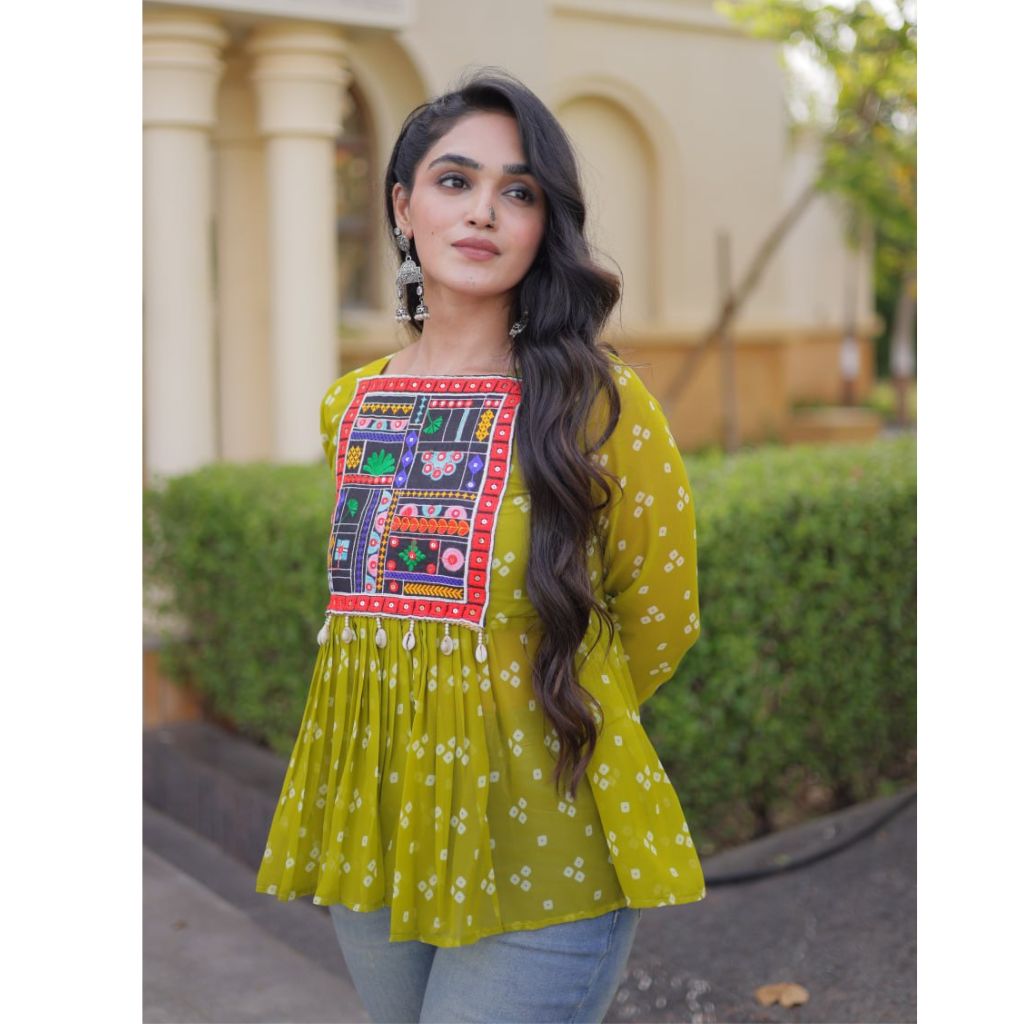 A woman wearing a yellow-green ethnic kurta with a colorful embroidered patch on the chest, paired with light blue jeans, standing outdoors in front of a beige building with columns.