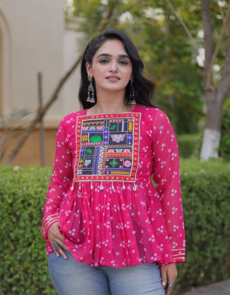 A woman wearing a bright pink long-sleeve kurta with a colorful embroidered patch on the chest, paired with light blue jeans, standing outdoors in front of a green hedge.