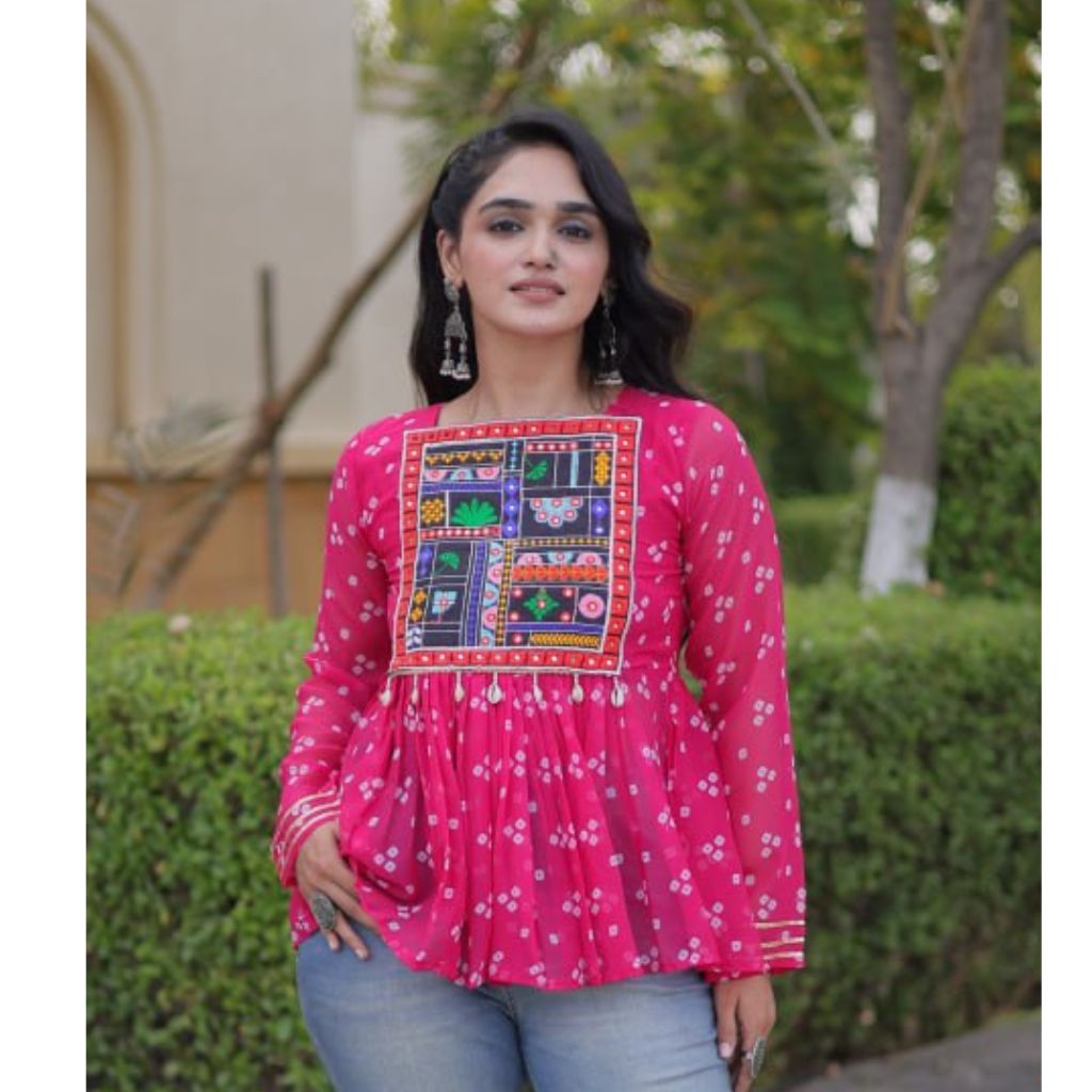A woman wearing a bright pink long-sleeve kurta with a colorful embroidered patch on the chest, paired with light blue jeans, standing outdoors in front of a green hedge.