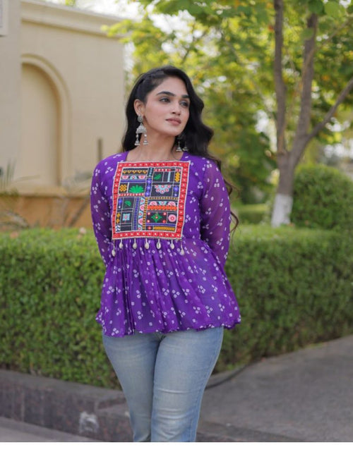 Load image into Gallery viewer, A woman wearing a purple long-sleeve top with a colorful embroidered patch on the chest and light blue jeans, standing outdoors in front of green hedges and a beige building.
