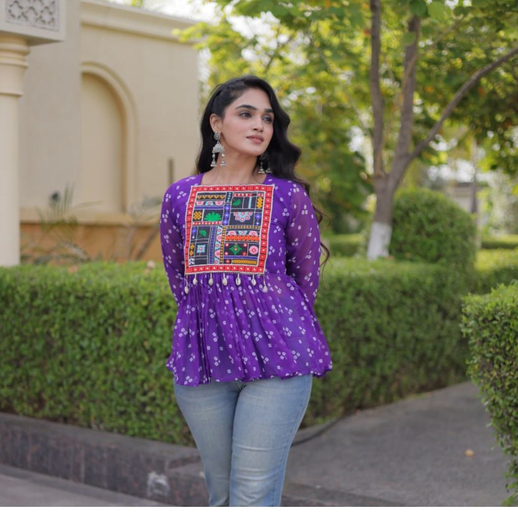 A woman wearing a purple long-sleeve top with a colorful embroidered patch on the chest and light blue jeans, standing outdoors in front of green hedges and a beige building.