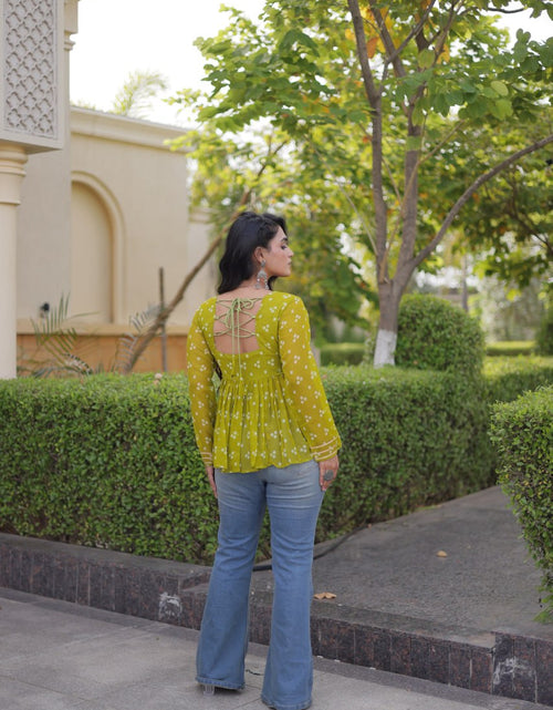 Load image into Gallery viewer, A woman stands outdoors on a paved walkway wearing a yellow-green long-sleeve blouse with a white floral print and blue flare jeans, photographed from the back against a background of green hedges and trees.
