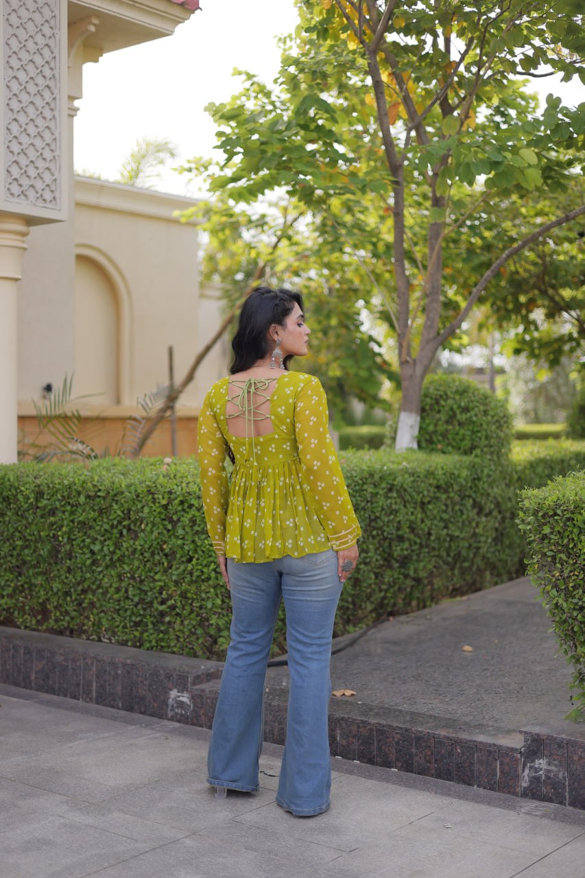 A woman stands outdoors on a paved walkway wearing a yellow-green long-sleeve blouse with a white floral print and blue flare jeans, photographed from the back against a background of green hedges and trees.
