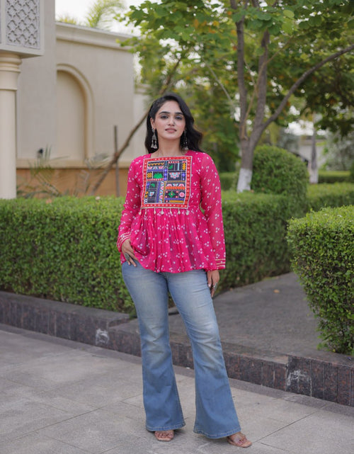 Load image into Gallery viewer, A woman stands outdoors on a paved walkway wearing a bright pink embroidered long-sleeve top with blue flared jeans. The background shows trimmed green hedges, trees, and a beige building.
