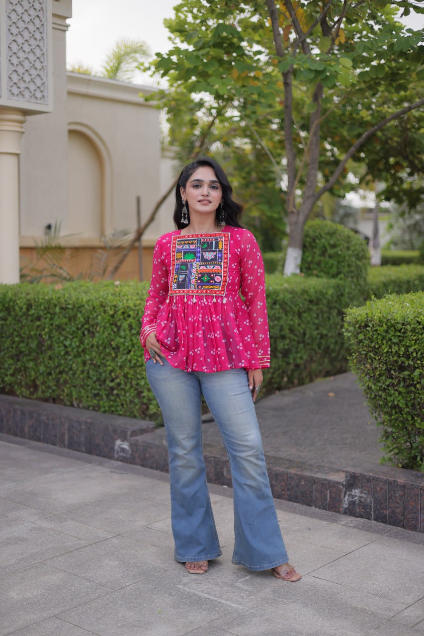 A woman stands outdoors on a paved walkway wearing a bright pink embroidered long-sleeve top with blue flared jeans. The background shows trimmed green hedges, trees, and a beige building.