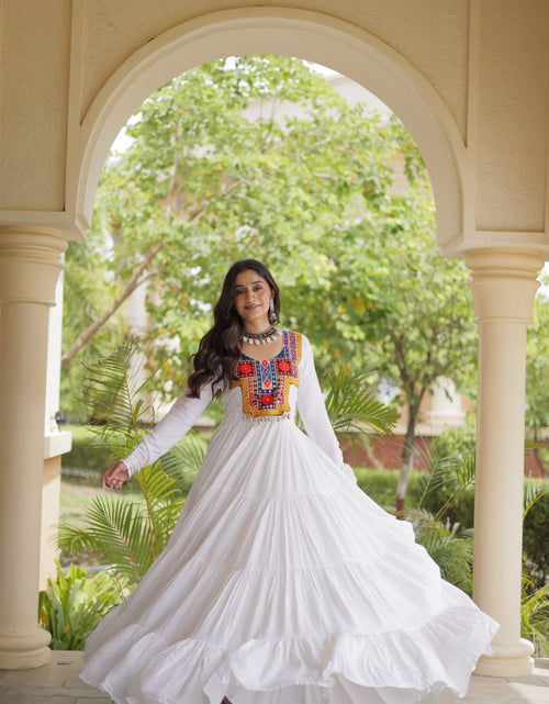 Load image into Gallery viewer, A woman wearing a long-sleeved white maxi dress with a colorful embroidered neckline stands in a courtyard framed by a beige archway, with green trees in the background.
