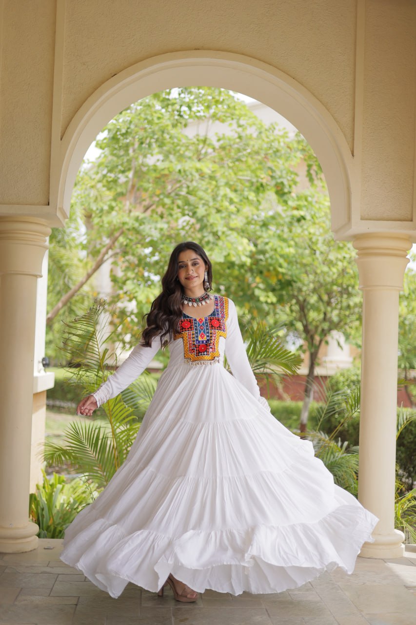 A woman wearing a long-sleeved white maxi dress with a colorful embroidered neckline stands in a courtyard framed by a beige archway, with green trees in the background.