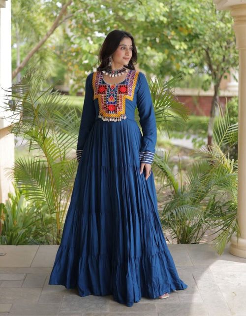 Load image into Gallery viewer, A woman models a long-sleeved navy blue maxi dress with a heavily embroidered neckline in red, orange, yellow, and white. She is standing on a stone patio in front of a classical building with cream pillars and green foliage in the background.
