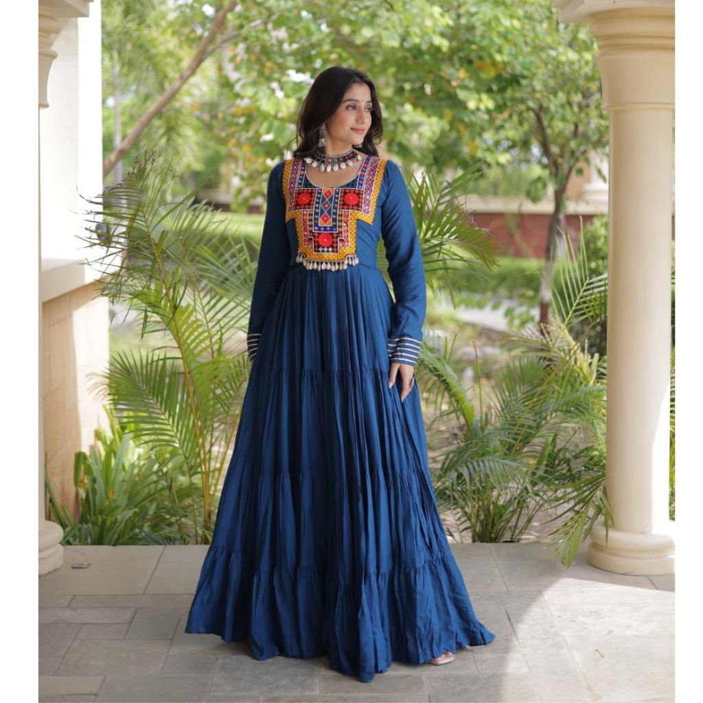 A woman models a long-sleeved navy blue maxi dress with a heavily embroidered neckline in red, orange, yellow, and white. She is standing on a stone patio in front of a classical building with cream pillars and green foliage in the background.