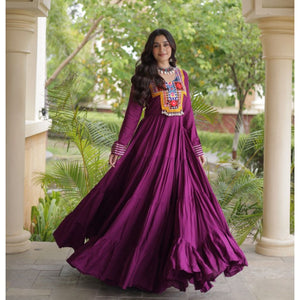 A woman wearing a long-sleeved magenta purple Anarkali gown with colorful geometric embroidery on the chest, standing in a garden courtyard framed by columns.