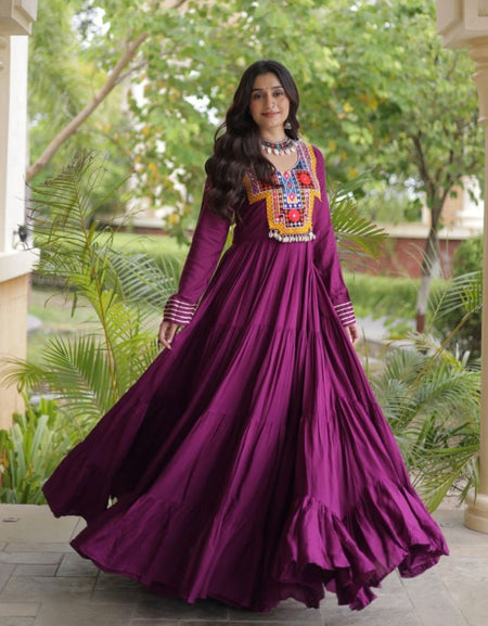 A woman wearing a long-sleeved magenta purple Anarkali gown with colorful geometric embroidery on the chest, standing in a garden courtyard framed by columns.