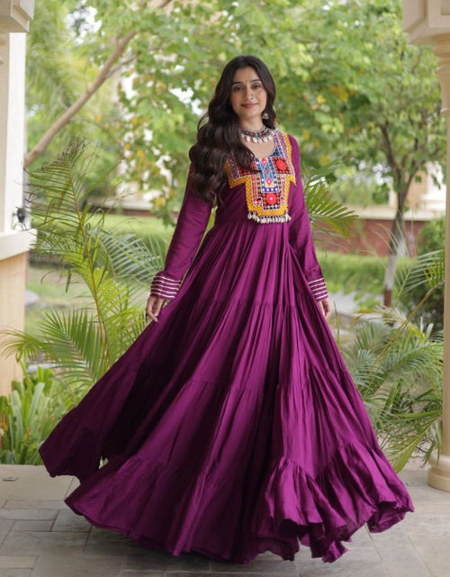 Load image into Gallery viewer, A woman wearing a long-sleeved magenta purple Anarkali gown with colorful geometric embroidery on the chest, standing in a garden courtyard framed by columns.
